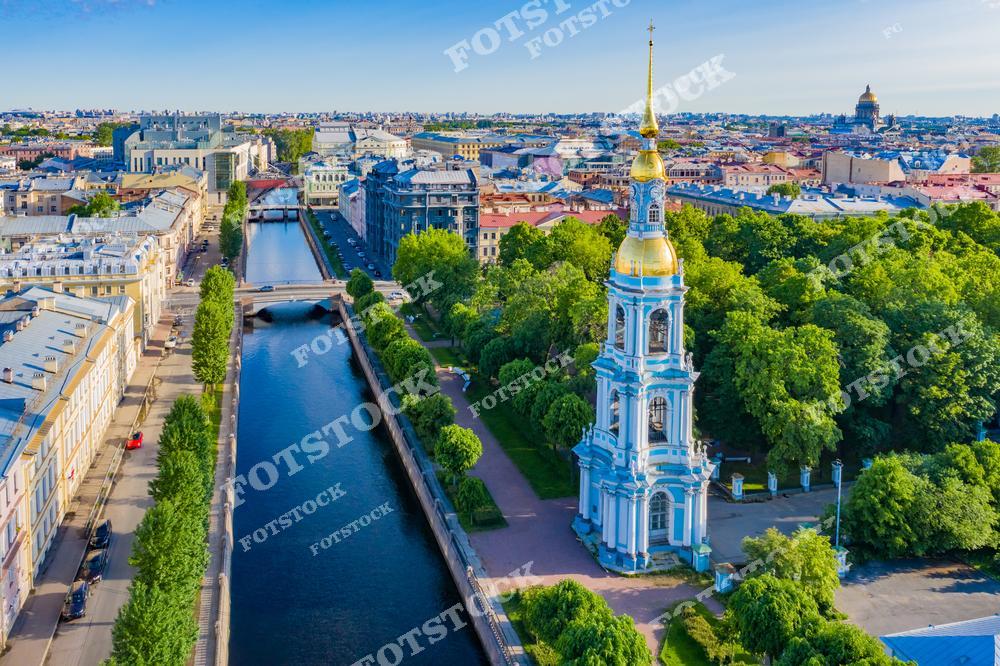 На переднем плане изображен Николо-Богоявленский морской собор с его характерной золотой колокольней. Собор находится рядом с каналом Грибоедова, который пересекает центральную часть Санкт-Петербурга. Деревья и зелень вокруг собора подчеркивают летнюю атмосферу. Вдалеке виднеется Исаакиевский собор и другие архитектурные достопримечательности города, создавая впечатляющую панораму.